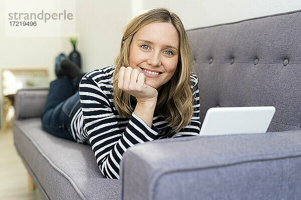 Lächelnde blonde Frau mit digitalem Tablet auf dem Sofa zu Hause liegend