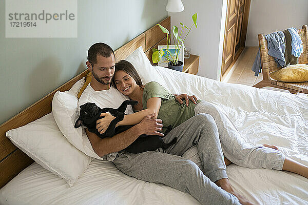 Verspieltes Paar  das einen Mops-Hund umarmt  während es zu Hause auf dem Bett liegt