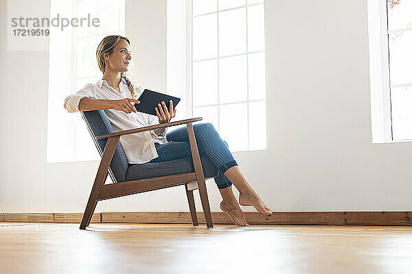Nachdenkliche Frau schaut weg  während sie ein digitales Tablet auf einem Stuhl am Fenster hält