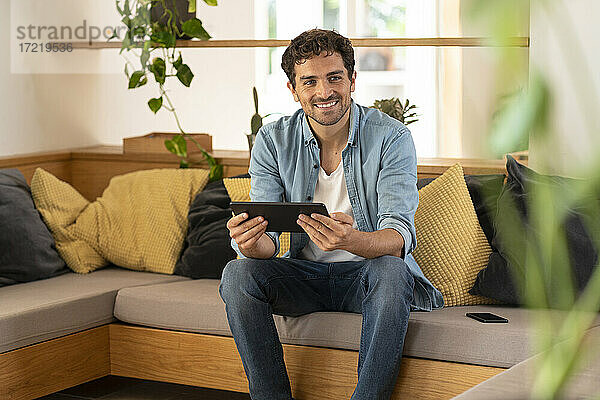 Lächelnder mittelgroßer erwachsener Mann  der wegschaut und ein digitales Tablet auf dem Sofa hält