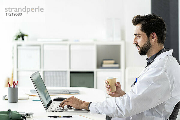 Männlicher Arzt  der einen Laptop benutzt  während er am Schreibtisch im Krankenhaus sitzt