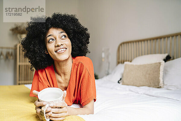 Nachdenkliche junge Frau mit Tasse  die zu Hause auf dem Bett liegt