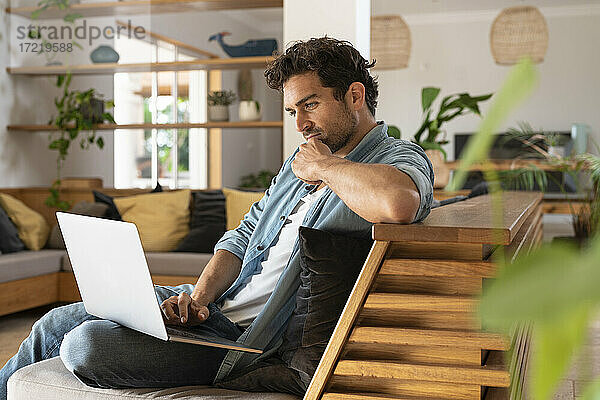 Konzentrierter Freiberufler  der zu Hause auf der Couch sitzt und einen Laptop benutzt