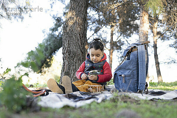 Unschuldiger Junge spielt mit einem Vergrößerungsglas an einem Rucksack in einem öffentlichen Park