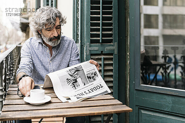 Mann trinkt Kaffee und liest Zeitung  während er am Tisch auf dem Balkon eines Hauses sitzt