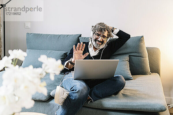 Gut aussehender Mann winkt während eines Videogesprächs auf dem Laptop  während er zu Hause auf dem Sofa sitzt