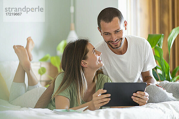 Junge Frau mit digitalem Tablet  die ihren Mann zu Hause auf dem Bett sitzend betrachtet