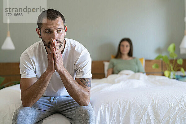 Ernster Mann auf dem Bett sitzend mit Freundin im Hintergrund zu Hause