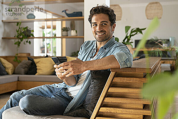 Lächelnder Mann  der wegschaut  während er sein Smartphone zu Hause in der Hand hält