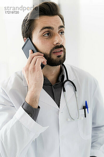 Gutaussehender männlicher Angestellter im Gesundheitswesen  der mit seinem Smartphone spricht und dabei wegschaut