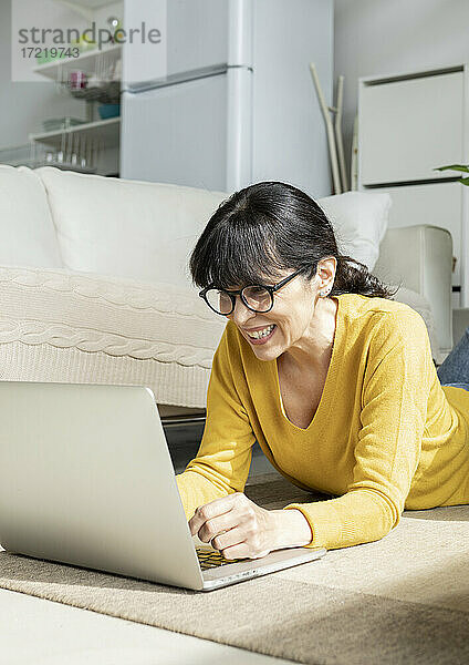 Lächelnde Frau  die einen Laptop benutzt  während sie zu Hause auf dem Boden liegt