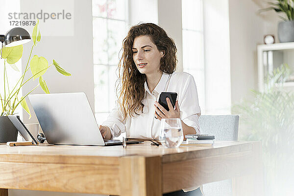 Schöne Unternehmerin  die einen Laptop benutzt und ein Smartphone am Schreibtisch im Home Office hält