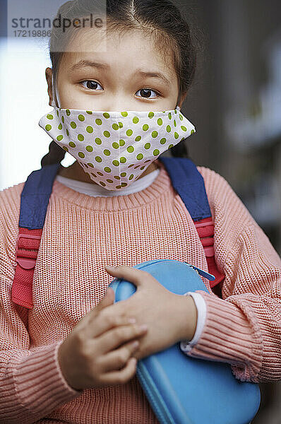Mädchen im Pullover mit Gesichtsschutzmaske hält Mäppchen während COVID-19