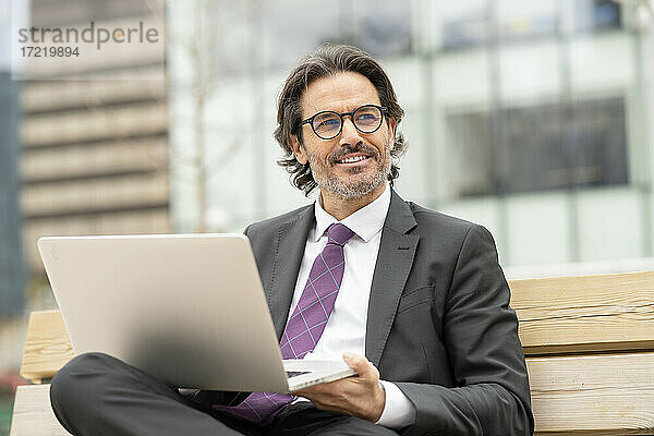 Lächelnder männlicher Unternehmer mit Laptop  der wegschaut  während er auf einer Bank sitzt