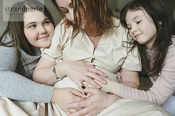 Aufgeregte schwangere Mutter und Töchter mit Händen auf dem Bauch sitzen zu Hause