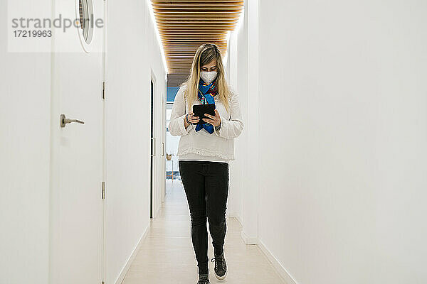 Weiblicher Patient mit Gesichtsmaske  der ein digitales Tablet benutzt  während er durch den Korridor eines medizinischen Gebäudes geht