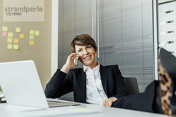 Glückliche Geschäftsfrau  die wegschaut  während sie am Schreibtisch mit ihrem Smartphone telefoniert