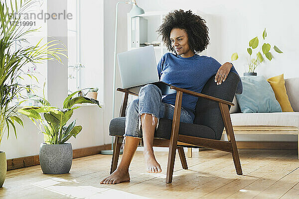 Lächelnde Frau  die einen Laptop benutzt  während sie zu Hause auf einem Sessel sitzt