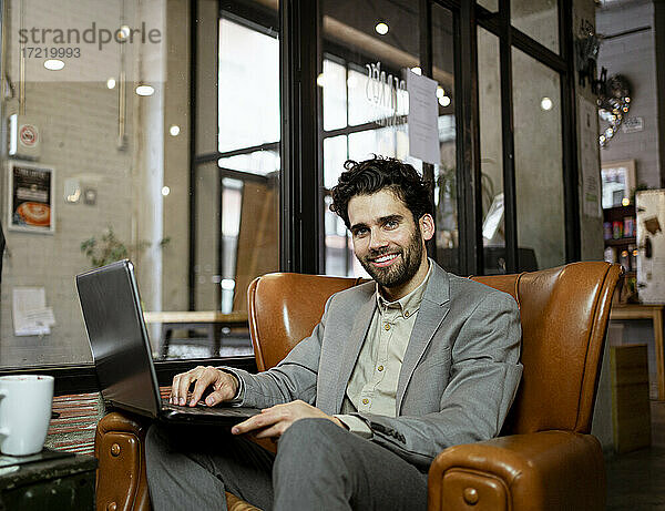 Lächelnde männliche Fachkraft auf einem Sessel sitzend mit Laptop in einem beleuchteten Café