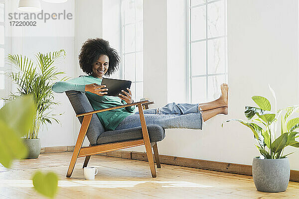 Lächelnde Frau  die ein digitales Tablet benutzt  während sie zu Hause auf einem Sessel sitzt
