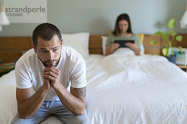 Nachdenklicher Mann mit Hand am Kinn  der mit seiner Freundin auf dem Bett sitzt und ein digitales Tablet zu Hause benutzt