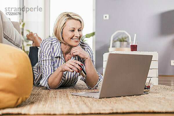 Lächelnde Frau mit Kreditkarte beim Online-Shopping  während sie zu Hause auf der Vorderseite liegt