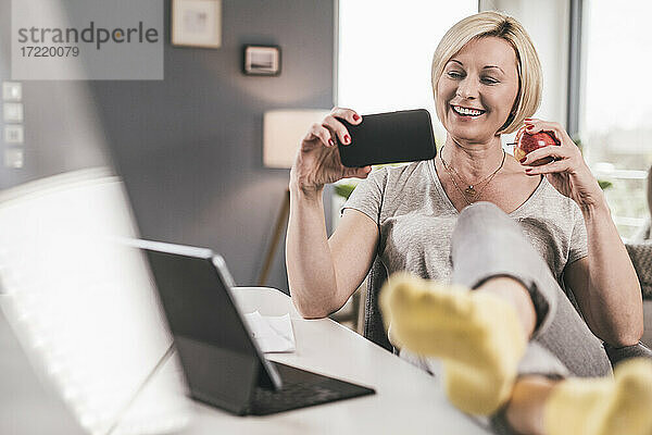 Lächelnde Frau mit Apfel  die zu Hause einen Film auf dem Mobiltelefon ansieht