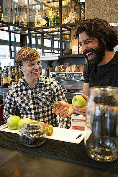 Männlicher Barkeeper lacht  während er frisches Obst von einem Auszubildenden an der Theke hält