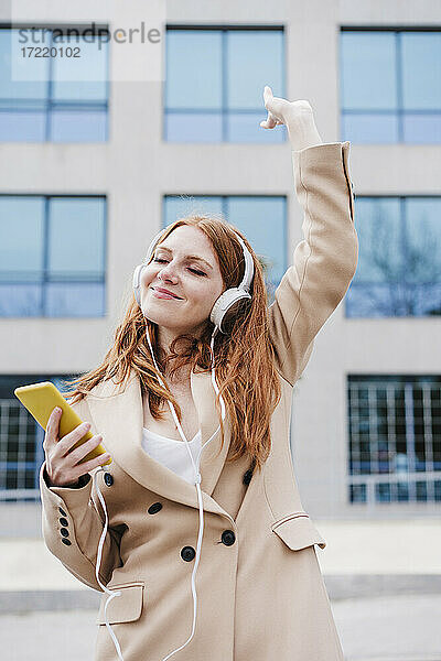 Lächelnde Frau mit Smartphone hört Musik über Kopfhörer und tanzt vor einem Gebäude