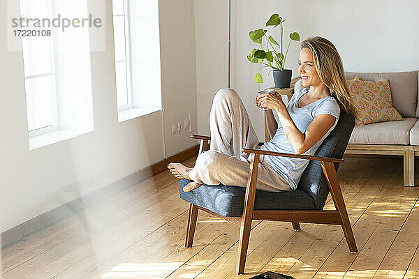Lächelnde Frau hält Kaffeetasse  während sie zu Hause auf einem Sessel sitzt