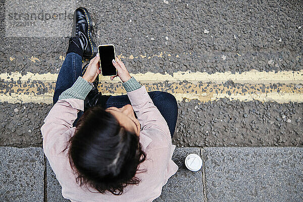 Junge Frau mit Mobiltelefon auf dem Fußweg sitzend