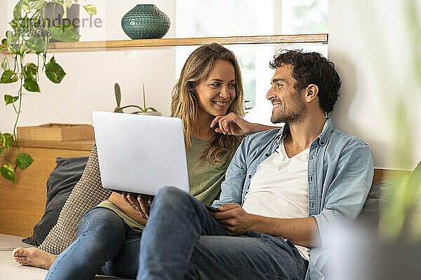 Lächelnder Mann mit Laptop  der seine Freundin anschaut  während er zu Hause auf der Couch sitzt