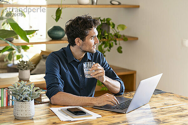 Nachdenklicher männlicher Freiberufler trinkt Wasser  während er mit seinem Laptop am Tisch sitzt