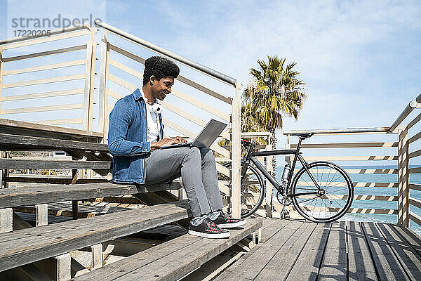 Lächelnder Mann  der einen Laptop benutzt  während er an einem sonnigen Tag auf den Stufen einer Fahrradpromenade sitzt