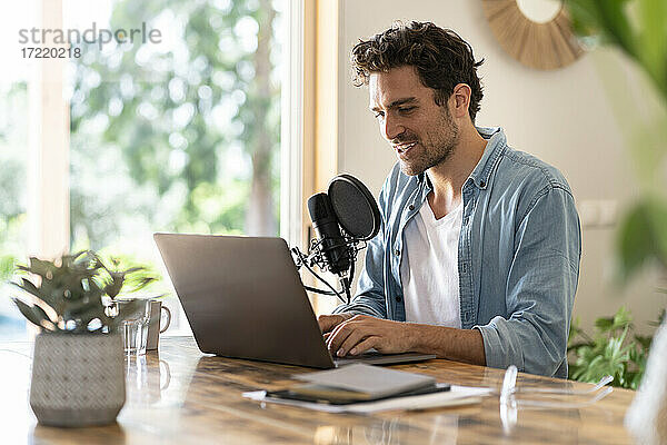 Männlicher Freiberufler  der vor einem Laptop in ein Mikrofon spricht  während er zu Hause einen Podcast aufnimmt
