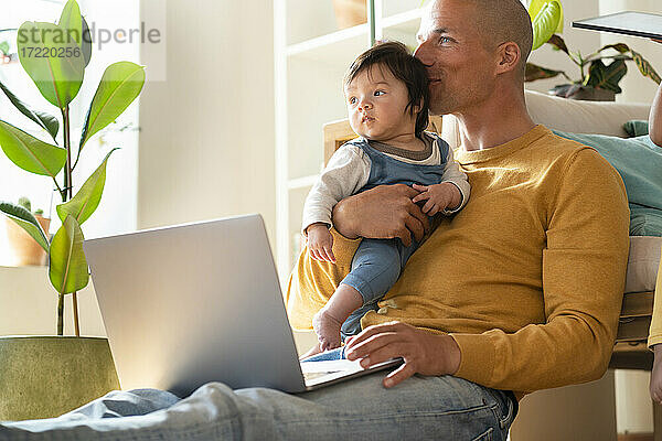 Vater mit Laptop  der sein Baby küsst  während er zu Hause sitzt