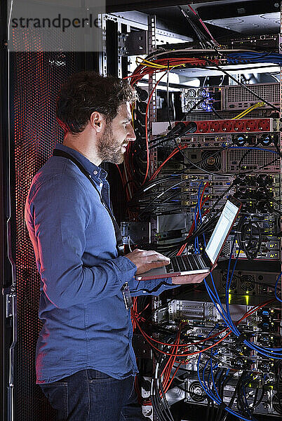 Lächelnder männlicher IT-Fachmann  der seinen Blick abwendet und einen Laptop im Serverraum hält