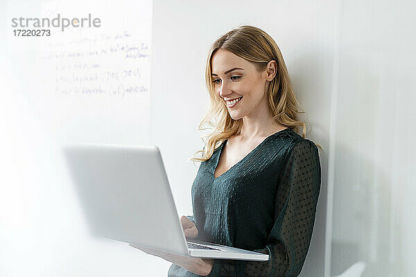 Lächelnde Geschäftsfrau  die einen Laptop benutzt und sich im Büro an die Wand lehnt