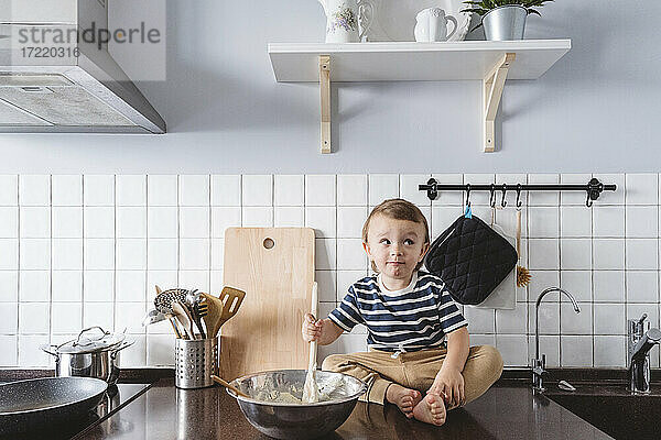Verspieltes Kleinkind mit Schöpfkelle und Schüssel  das wegschaut  während es auf der Küchentheke zu Hause sitzt