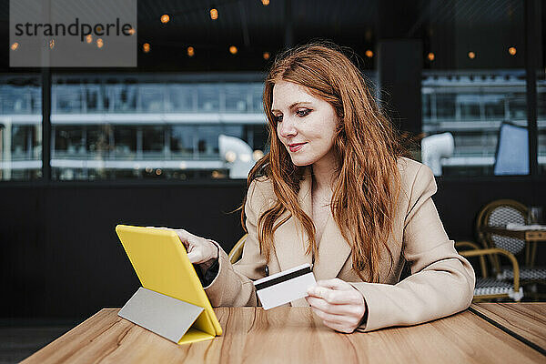 Rothaarige Frau  die eine Kreditkarte hält und ein digitales Tablet in einem Café benutzt