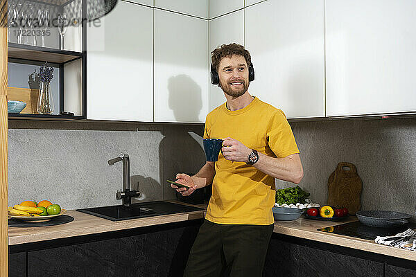 Mann hört Musik und hält Handy und Kaffeetasse in der Küche zu Hause