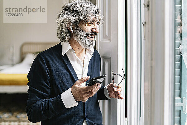 Lächelnder älterer Mann mit In-Ear-Kopfhörern  der in der Nähe des Fensters zu Hause mit seinem Smartphone telefoniert