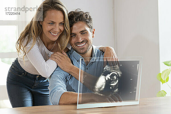 Verliebtes Paar betrachtet Ultraschallbild auf transparentem Bildschirm zu Hause