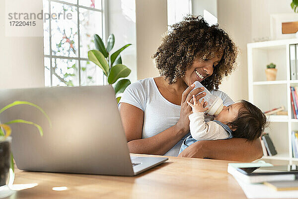 Lächelnde Mutter  die ihr Baby mit Milch füttert  während sie im Büro zu Hause am Laptop sitzt