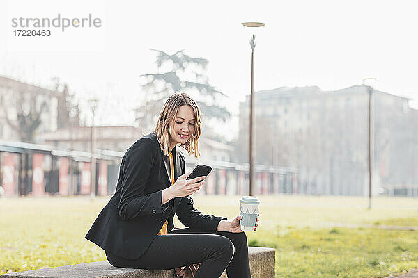 Mittlere erwachsene Geschäftsfrau mit Kaffee-Einwegbecher und Mobiltelefon im Park