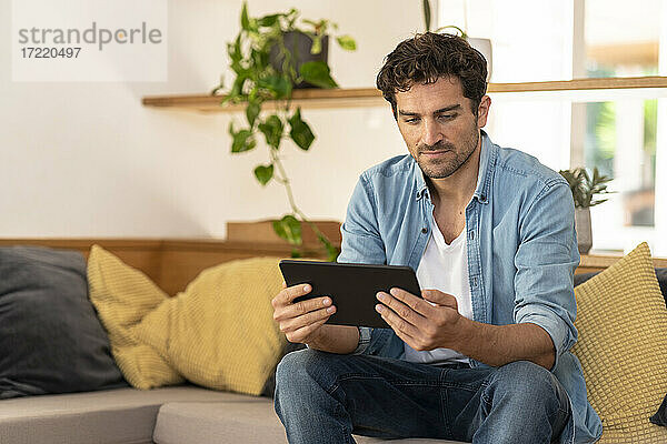 Ernster Mann  der ein digitales Tablet benutzt  während er zu Hause auf der Couch sitzt