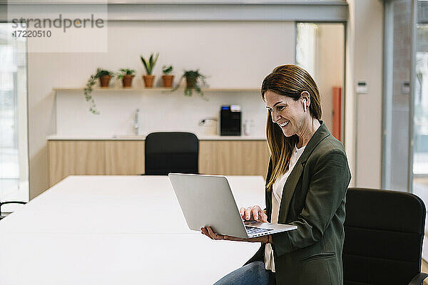 Lächelnde Unternehmerin mit In-Ear-Kopfhörern  die einen Laptop benutzt und sich auf einen Tisch im Büro stützt