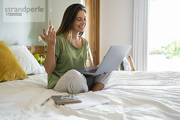 Junge Frau gestikuliert während eines Videoanrufs auf dem Laptop  während sie zu Hause auf dem Bett sitzt