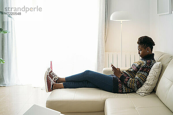 Junge Frau benutzt ihr Smartphone  während sie auf dem Sofa im Wohnzimmer sitzt