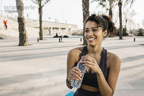 Lächelnde Sportlerin  die mit einer Wasserflasche in der Hand auf dem Fußweg wegschaut